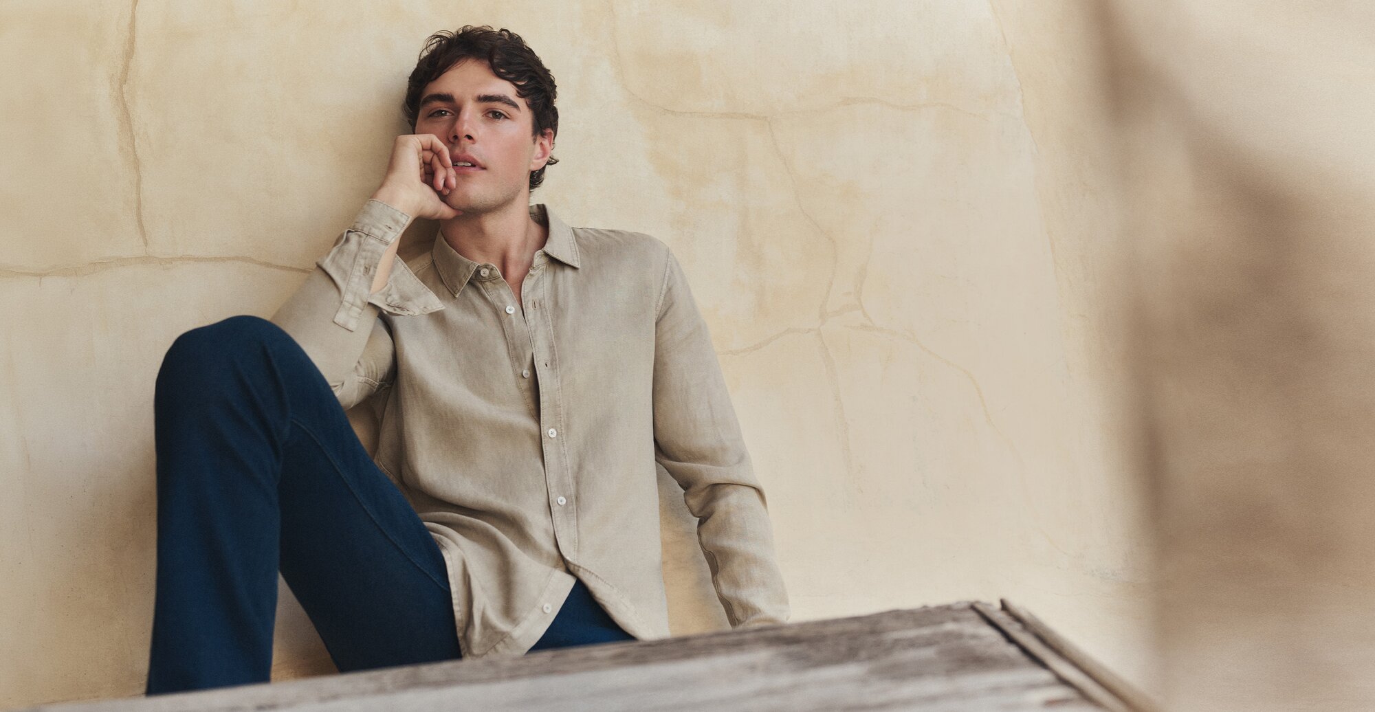 Man lounging in a taupe long-sleeve button-front shirt and dark wash jeans.