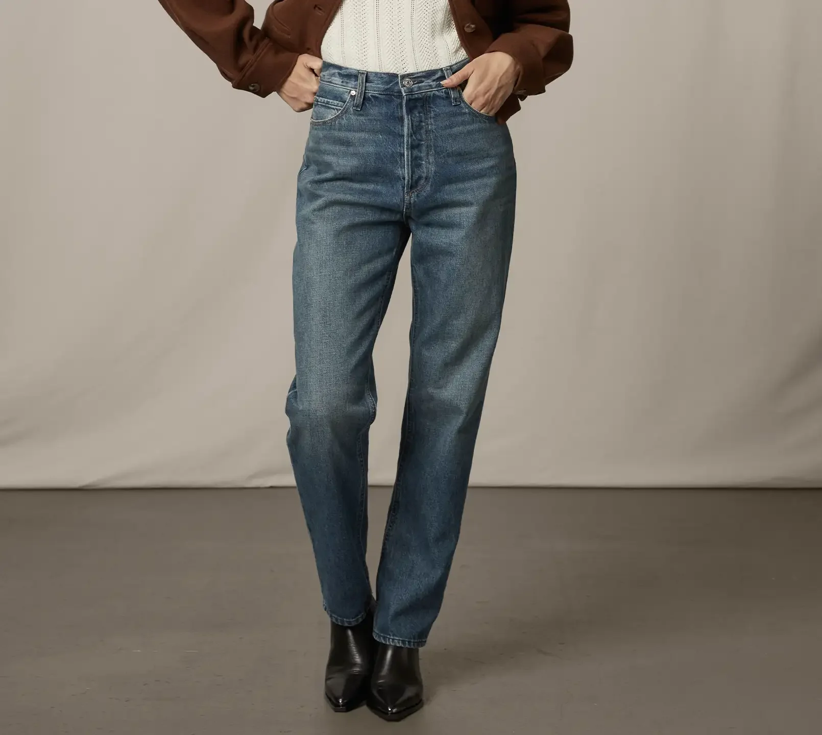 Woman standing wearing dark wash jeans, a white sweater top, brown jacket, and black boots