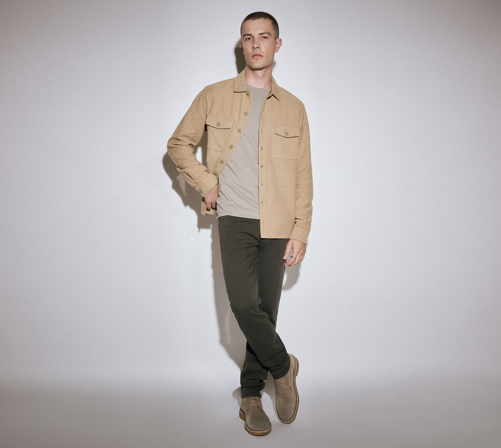 Man wearing a tan overshirt over a grey tee, dark grey jeans, and grey boots