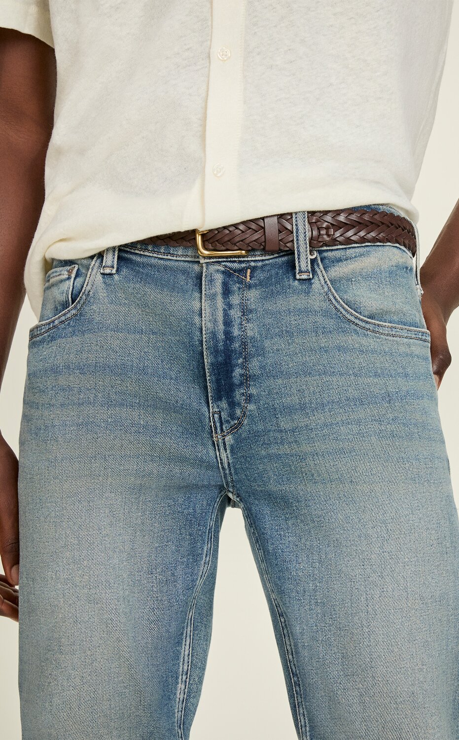 Close-up of a model wearing light-wash slim-fit jeans paired with a cream short-sleeve shirt and a brown woven leather belt, highlighting the denim fit and wash.