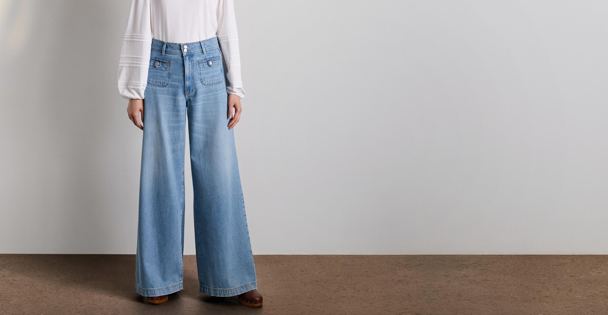 Woman wearing a long sleeve white top tucked into light wash wide leg jeans and auburn clogs