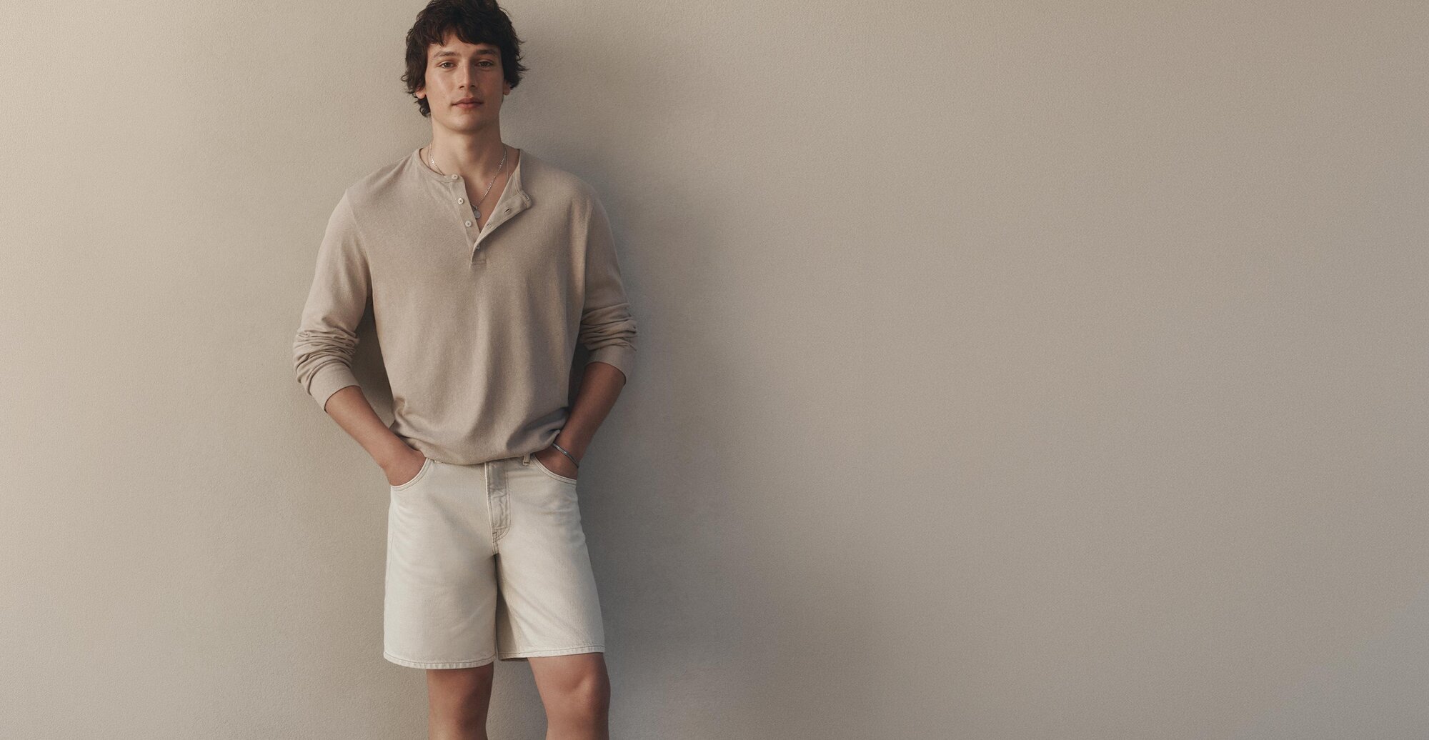 Man wearing a long-sleeve henley shirt and light shorts, standing against a neutral wall.