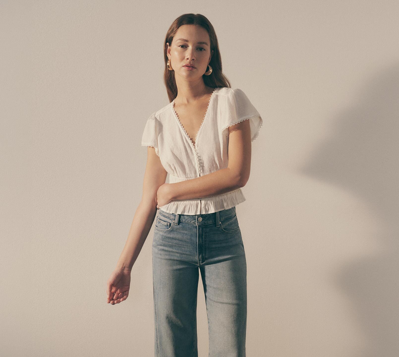 Woman wearing a white belled sleeve top with lace trim detail and light wash denim jeans all by PAIGE.