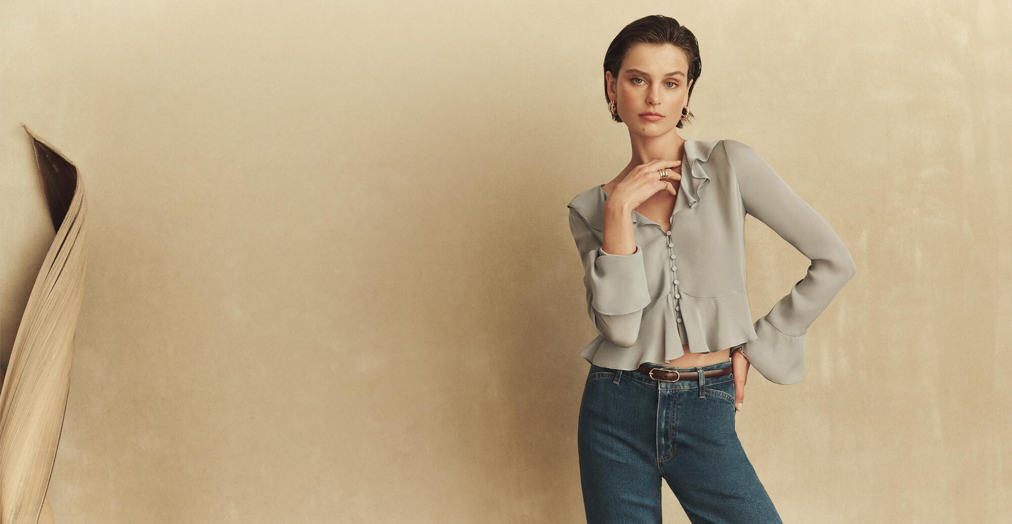 A woman wearing a light blue button-up long sleeve blouse, medium blue wash denim jeans and a thin brown leather belt with gold buckle detail standing against a beige wall.