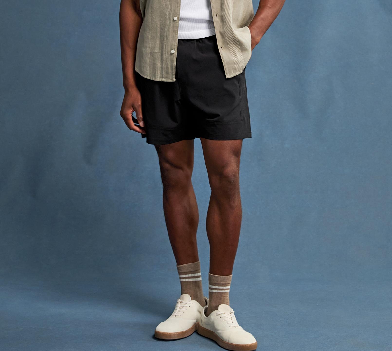 Man wearing a green button up shirt over a white tank, black shorts, and ivory sneakers