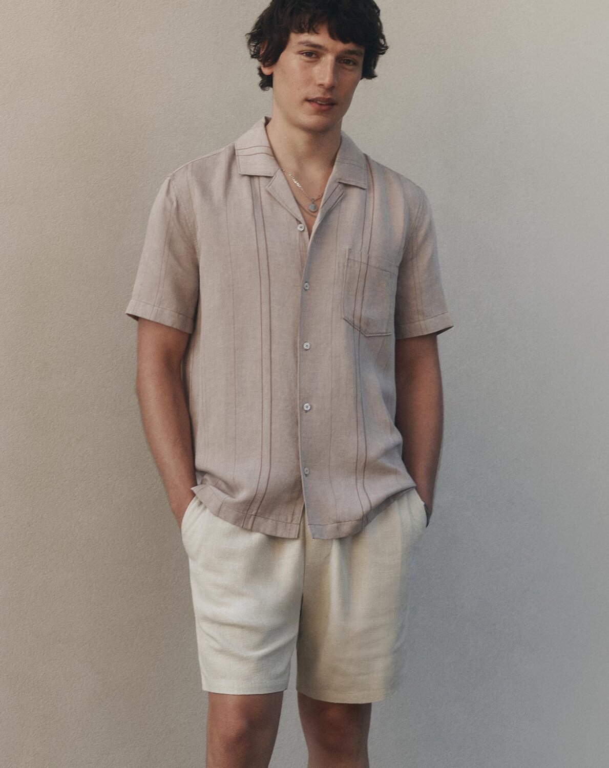 Man wearing a short-sleeve button-up shirt and light shorts, standing against a neutral wall.