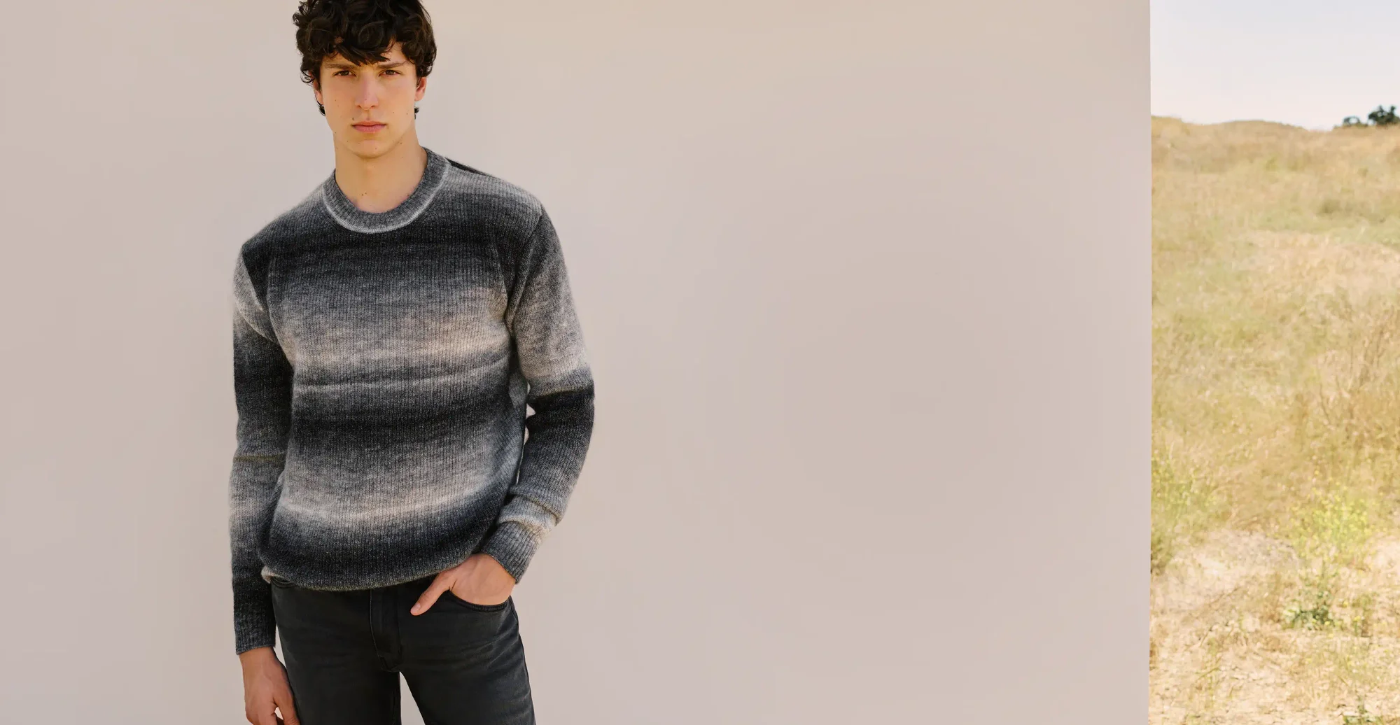 Man standing wearing a grey and black gradient striped sweater and dark wash jeans