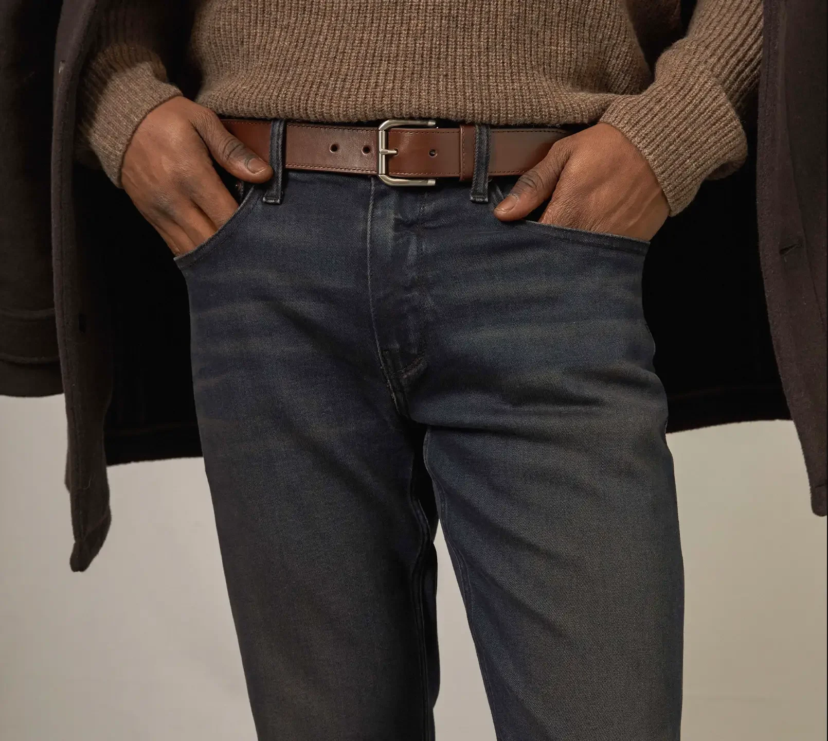 Man standing wearing dark wash jeans, a brown belt, a brown sweater, and a brown coat