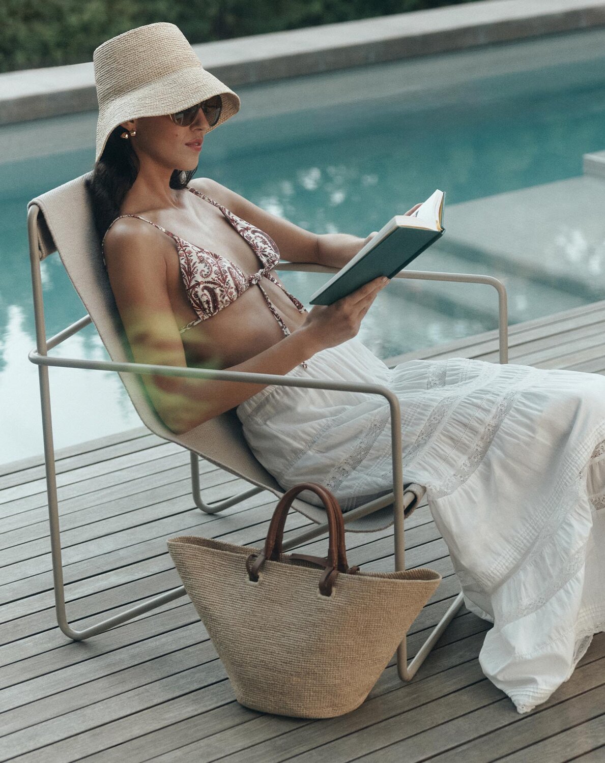 Woman relaxing poolside in a bikini top and white skirt, wearing a straw hat and reading a book.