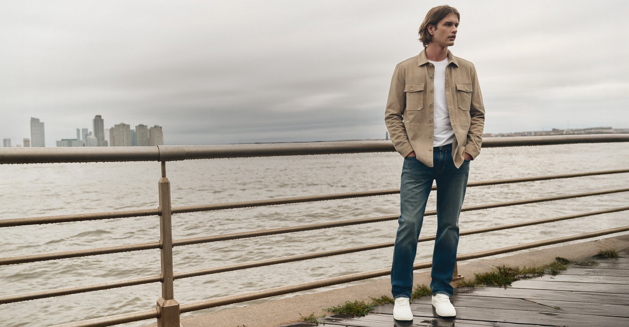 Man wearing a light grey suede jacket over a white tee, dark wash jeans, and white sneakers