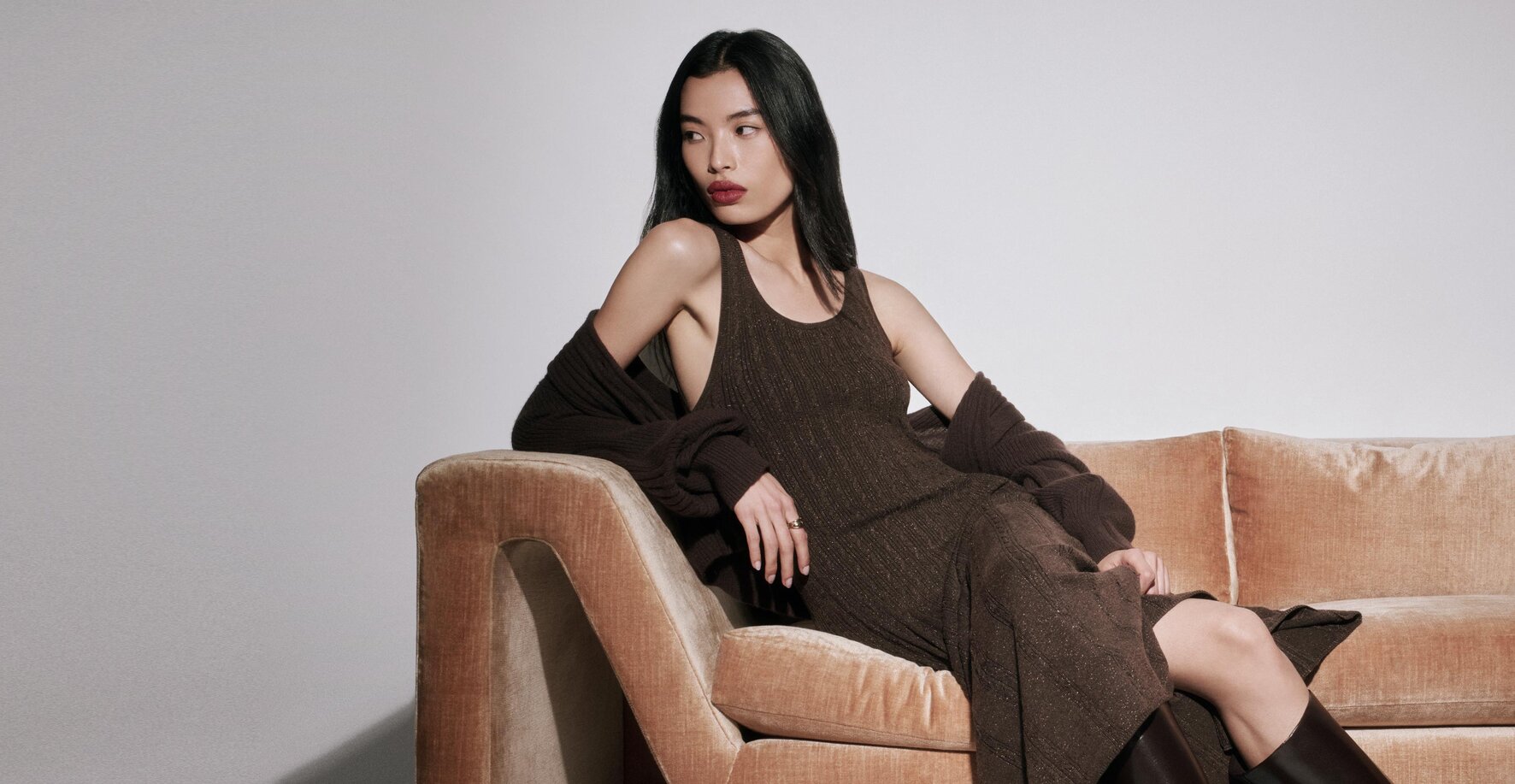 Woman sitting on a couch wearing a long brown sparkle dress with a brown cardigan and tall brown boots