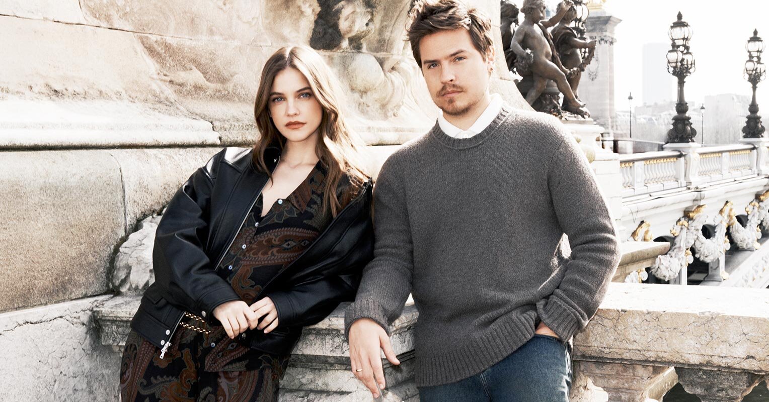 An image of Barbara Palvin and Dylan Sprouse for the Fall 2026 PAIGE campaign. Barbara wears a maroon and black paisley top and matching wide leg pants paired with a black oversized jacket in faux leather and gold chain belt. Dylan wears a grey sweater, white shirt and blue denim jeans.