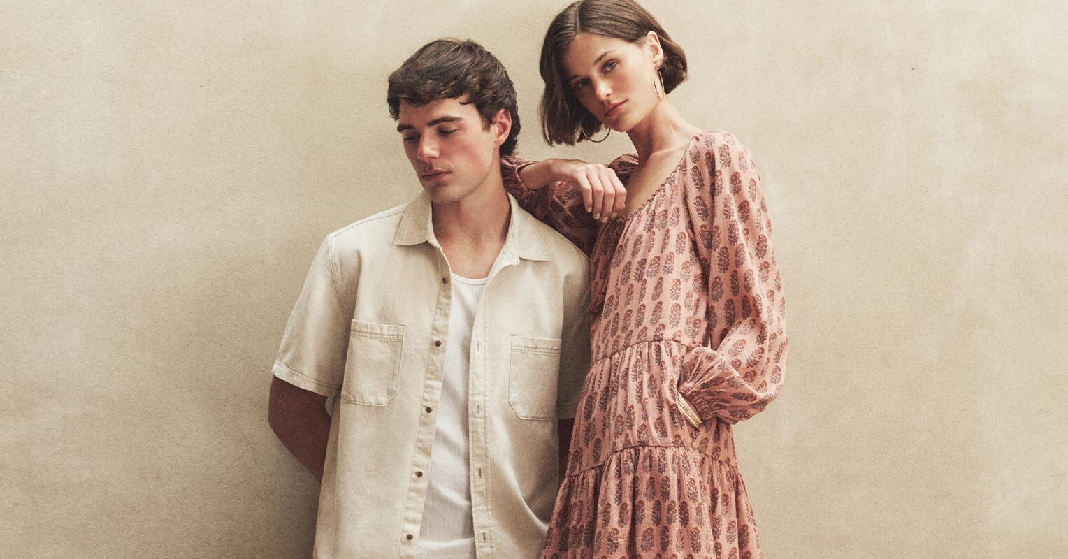 Man wearing a biege short sleeve button up denim shirt with white ribbed tank standing next to a woman wearing a pink long sleeve mini-dress with embroidered pattern detail.