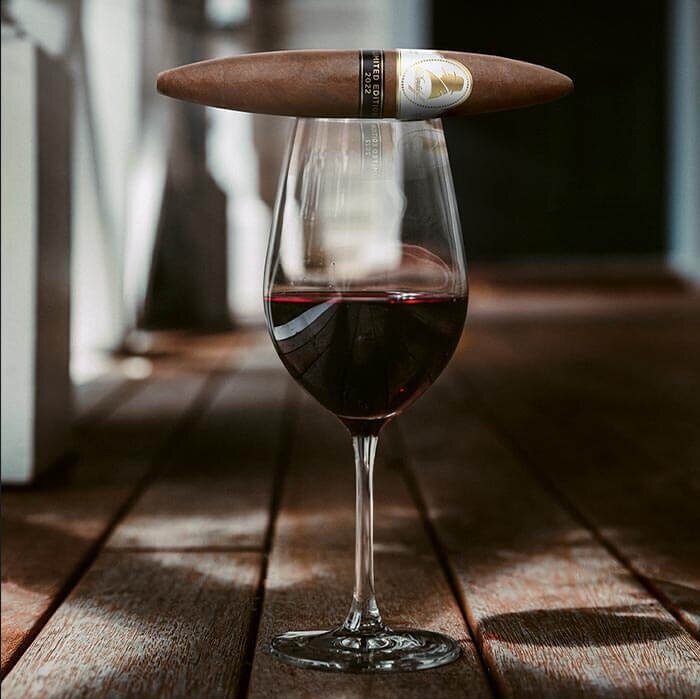 A luxurious cigar resting on the rim of a wine glass filled with red wine, set against a wooden floor background.