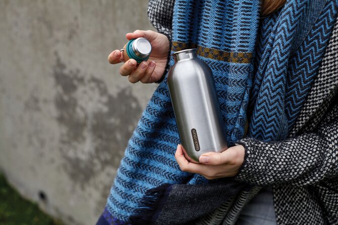 Stilren vannflaske i rustfritt stål med avtakbart lokk, holdt av en person iført et varmt skjerf. Flasken er ideell for turer og hverdagsbruk. Elegant design kombinert med funksjonalitet.