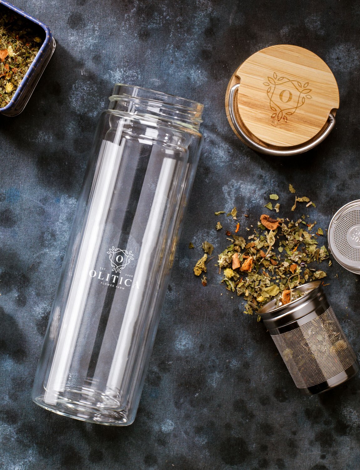 Eksklusiv glassflaske med te-infuser og bambuslokk, omgitt av løse teblader og en metallbeholder. Flasken har et minimalistisk design med gravert logo og gir en stilren følelse av bærekraft og velvære. Perfekt for teelskere på farten.