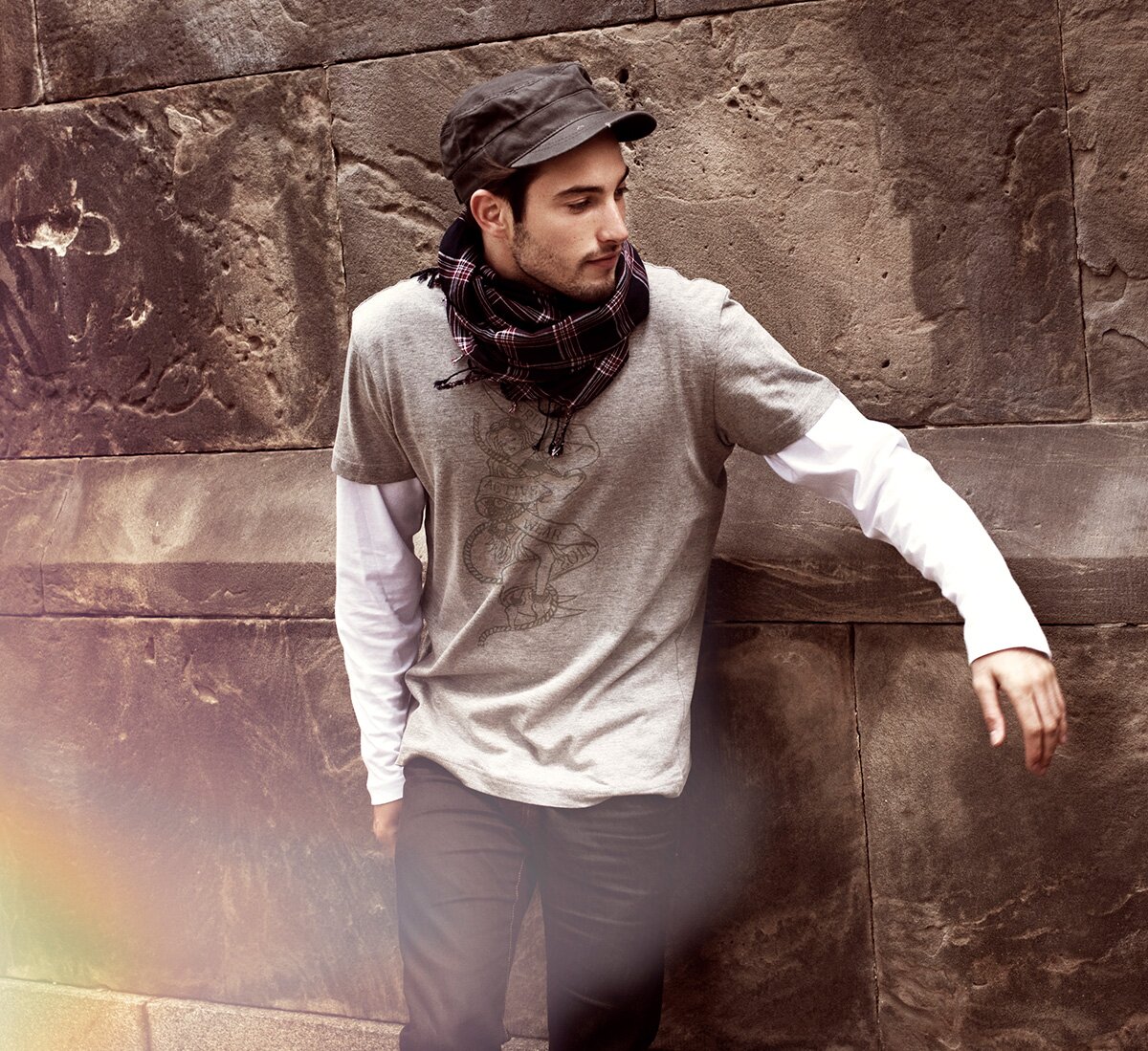 Mann i grå t-skjorte med grafisk trykk, hvit langermet genser og rutete skjerf fra Printer, står mot en murvegg.
