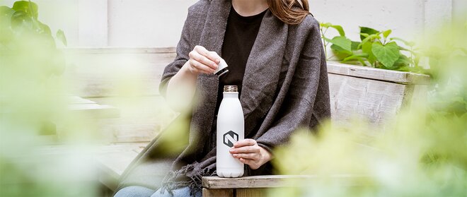 Kvinne iført grå sjal holder en hvit termoflaske med logo. Elegant og funksjonell firmagave for varm og kald drikke.