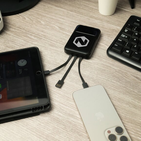 Sort powerbank med integrerte ladekabler og gravert firmalogo, koblet til mobil og nettbrett. Praktisk og stilren firmagave for lading av flere enheter samtidig.