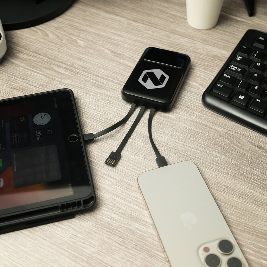 Sort powerbank med integrerte ladekabler og gravert firmalogo, koblet til mobil og nettbrett. Praktisk og stilren firmagave for lading av flere enheter samtidig.
