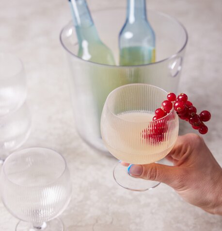 Stemningsfullt bilde av et rillet Sagaform-glass fylt med drink, dekorert med ripsbær, og en isbøtte med flasker i bakgrunnen.