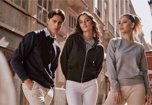 Tre unge voksne i Roly-gensere og hettejakke i nøytrale farger, stående i en bygate.