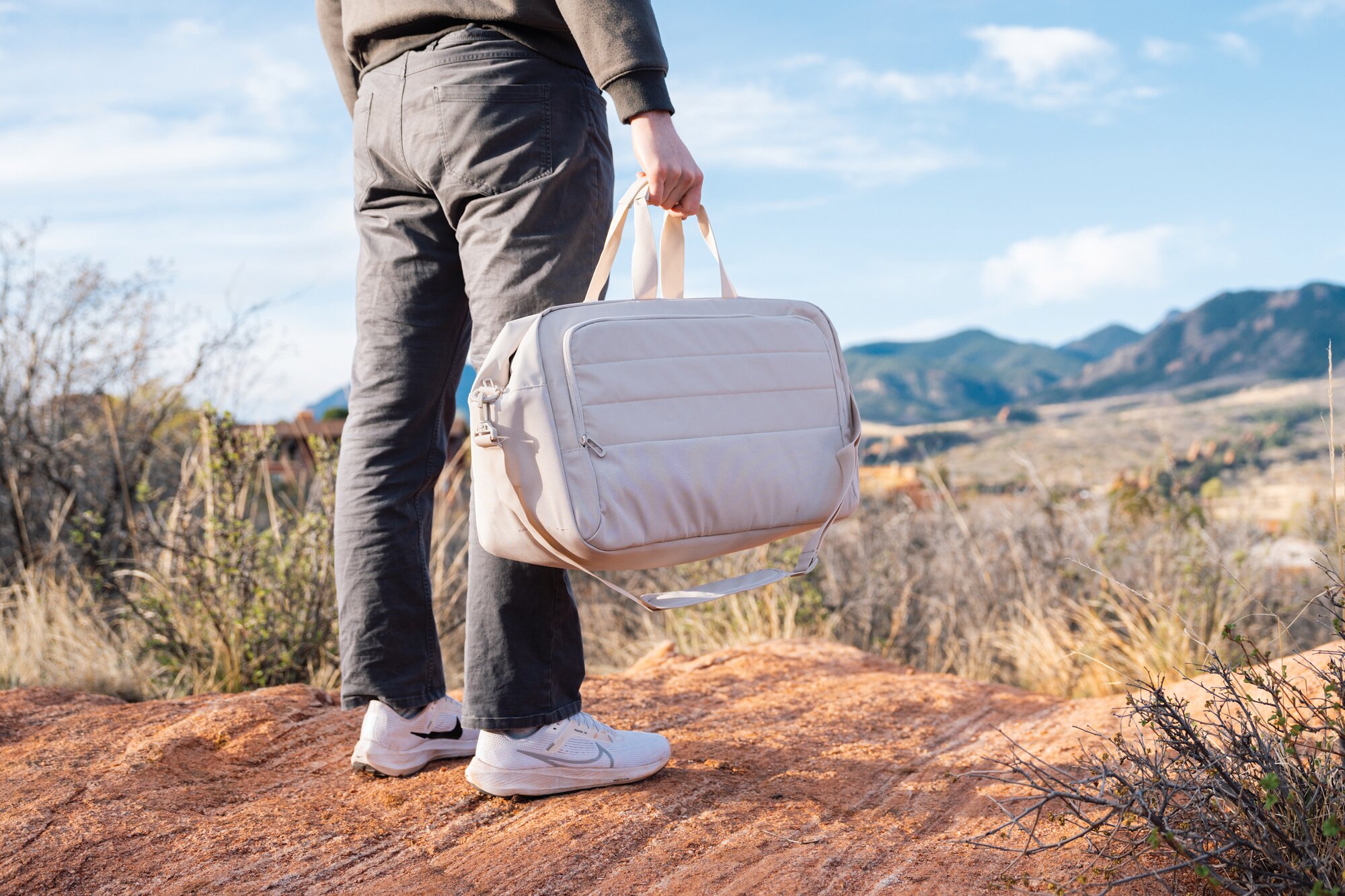 En person står i et fjellandskap med en beige XD Xclusive-bag i hånden.