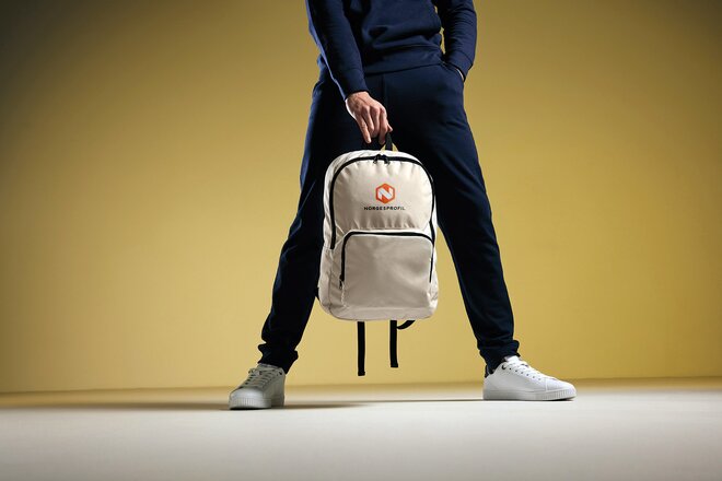 Person i mørk joggedress og hvite sneakers holder en beige ryggsekk med trykket firmalogo mot en gul bakgrunn. Trendy og funksjonell profileringssekk for daglig bruk, jobb og events.