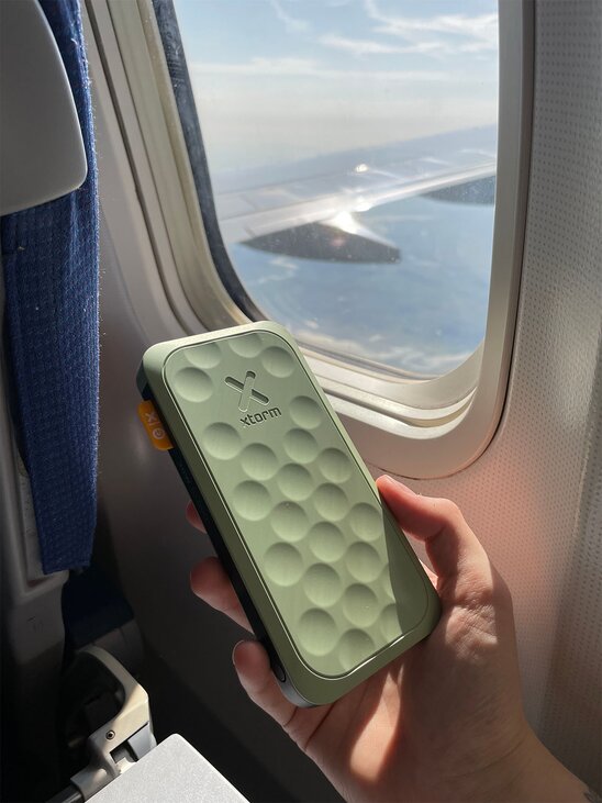 En grønn Xtorm-powerbank holdes ved et flyvindu under en flytur.