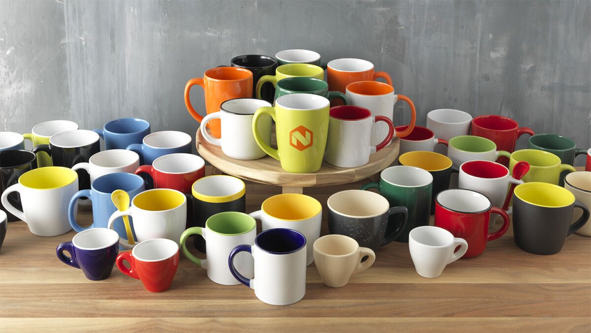 Fargerike krus i ulike størrelser og design plassert på et trebord, med en grå betongvegg i bakgrunnen. Perfekt for profilering med logo.