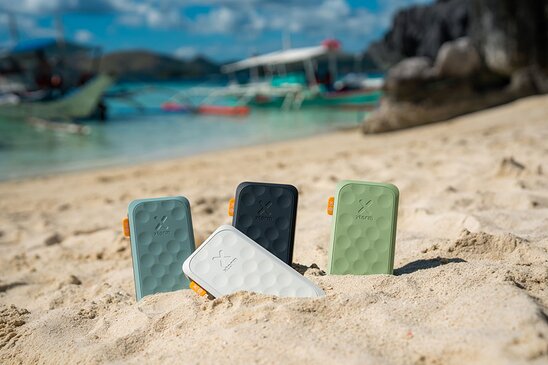 Fire Xtorm-powerbanks i ulike farger stikker opp av sanden på en tropisk strand.