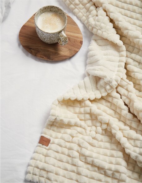 Mykt, kremfarget Vinga-pledd på en seng, med en kaffekopp på et rundt trebrett.