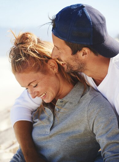 Smilende par på stranden, kvinnen i grå piké og mannen i hvit t-skjorte og caps fra Printer.