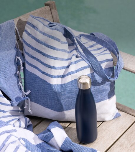 Sagaform strandveske i blått og hvitt med en matchende Sagaform termosflaske i mørk blå, plassert på en trebenk ved et svømmebasseng. Perfekt for strand og piknik.