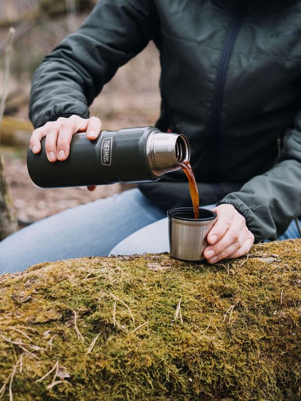 Nærbilde av en grønn Thermos-flaske mens kaffe helles i den medfølgende koppen, i skogsmiljø.