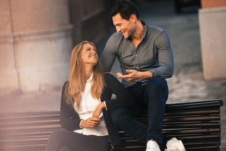 En kvinne i hvit skjorte og sort jakke sitter smilende ved siden av en mann i mørk skjorte på en benk.