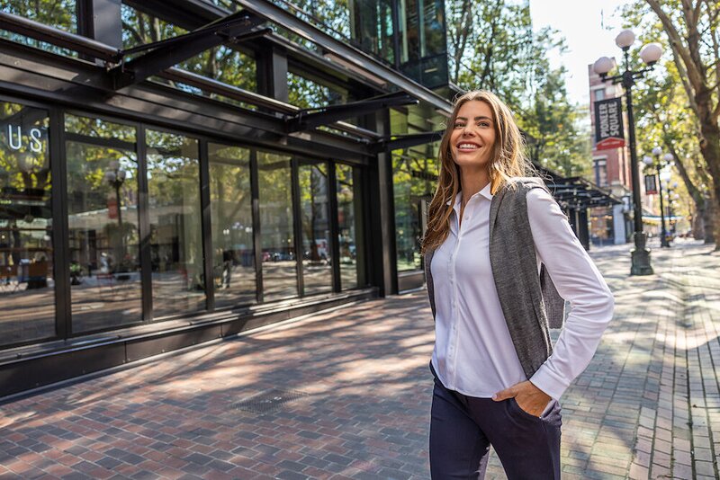 Kvinne i hvit skjorte og grå vest fra J. Harvest & Frost, går smilende gjennom en bygate.