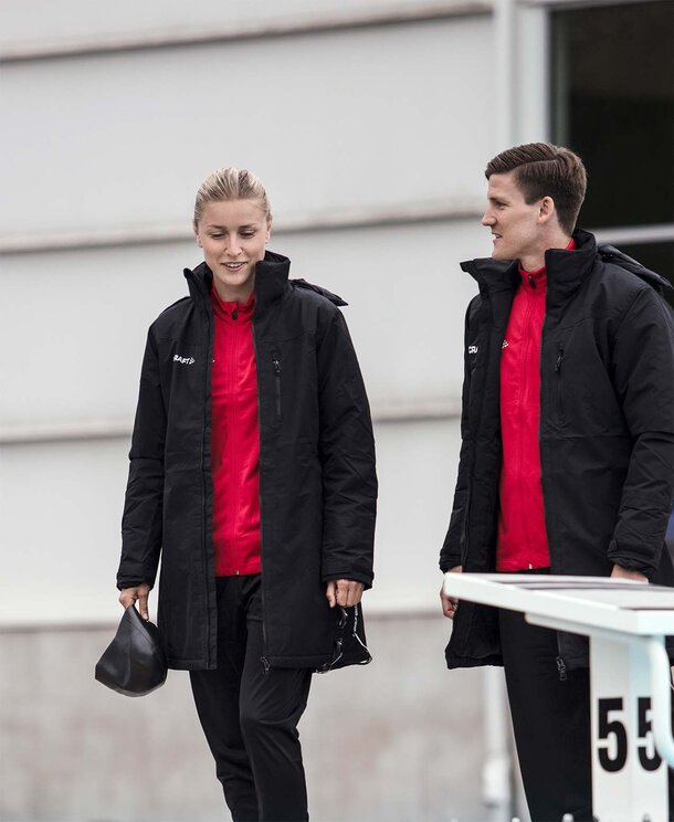En kvinne og en mann i svarte Craft Teamwear-parkaser, på en sportsarena.