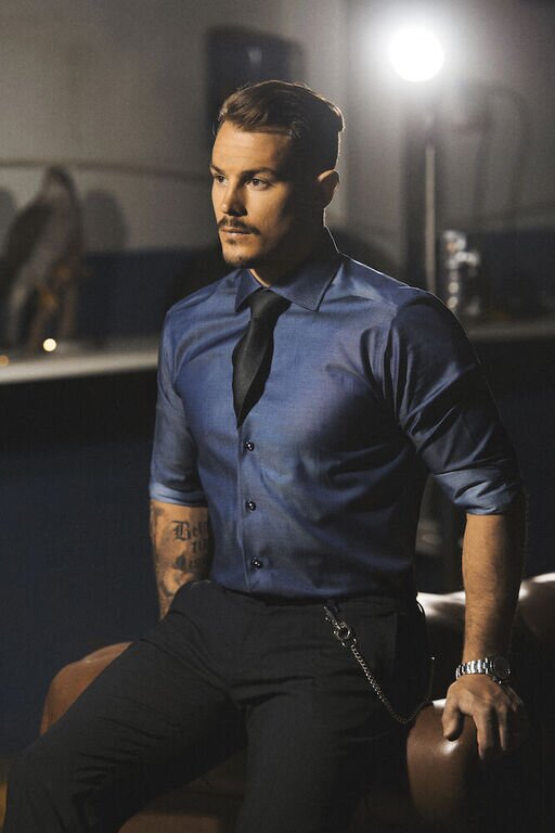 Mann i en skinnende blå J. Harvest & Frost-skjorte og sort slips, sittende på en stol i et mørkt rom.