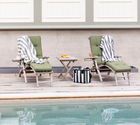 Sagaform solsenger med grønne puter og grå- og hvitstripede Sagaform håndklær, plassert ved et basseng, med en liten trekrakk mellom dem som holder Sagaform glass med drikke. Perfekt for avslapning utendørs.