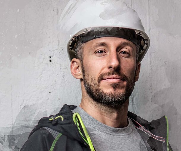 «Mann iført hvit vernehjelm og mørk Jobman-jakke, vendt mot kamera foran en grå bakgrunn.