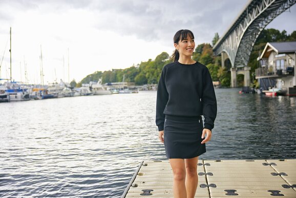 Kvinne iført svart collegegenser og matchende skjørt, stående ved vannkanten – en elegant og allsidig profilgenser for bedrifter og events.