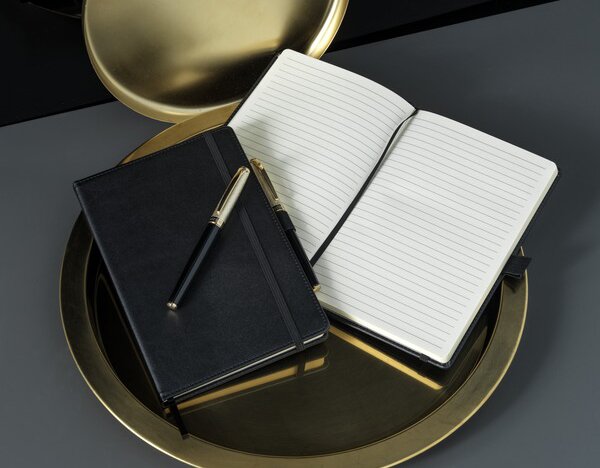 Elegant oppsett av en svart JournalBooks-notatbok med elastisk bånd, plassert på et gullfarget fat. En åpen notatbok med linjerte sider og en svart penn ligger ved siden av.