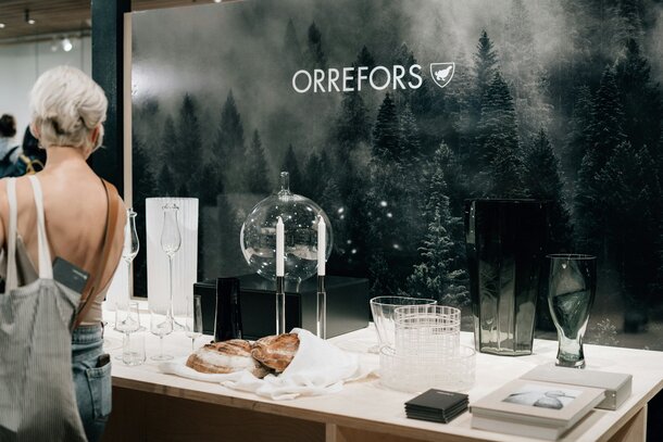 «Utstillingsbord med ulike Orrefors-glass og -vaser foran en mørk skogbakgrunn, med Orrefors-logoen over.