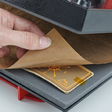 Påføring av varmeoverført stoffmerke med broderte detaljer. En hånd trekker tilbake beskyttelsespapir fra et brunbeige stoffmerke med gult kompassmotiv etter varmepressing. Motivet er trykket, men har en brodert effekt.