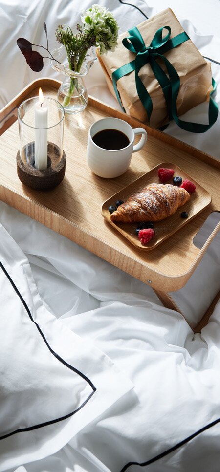 Frokost på sengen med trebrett fra Sagaform, inkludert kaffe, croissant med bær, en gave med grønt bånd og en tent lyslykt.