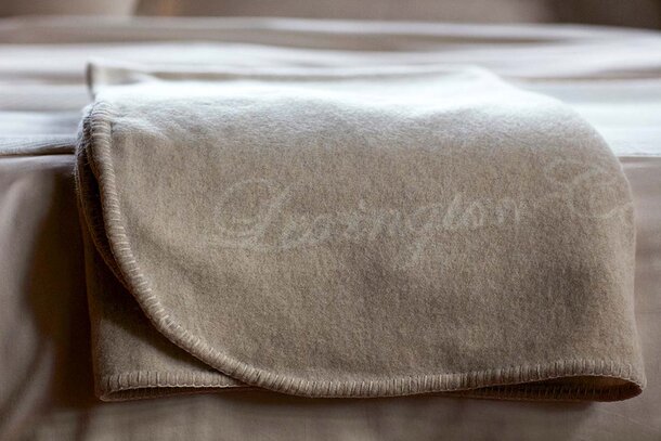 Nærbilde av et mykt, beige Lexington-pledd med brodert logo, liggende på en oppredd seng.