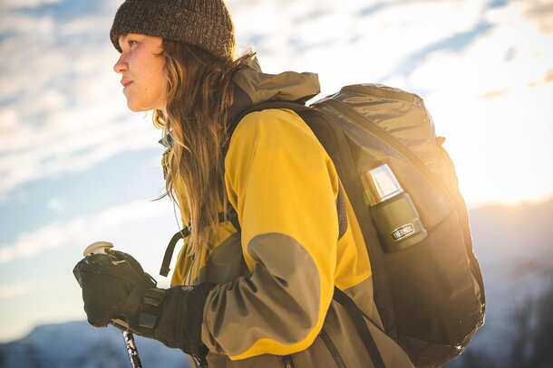 Kvinne i gul og grå jakke med en Thermos-flaske festet til ryggsekken, klar for tur i fjellet.