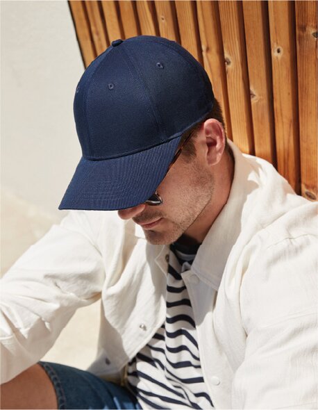 Mann med mørkeblå caps fra Beechfield, sittende mot en trevegg