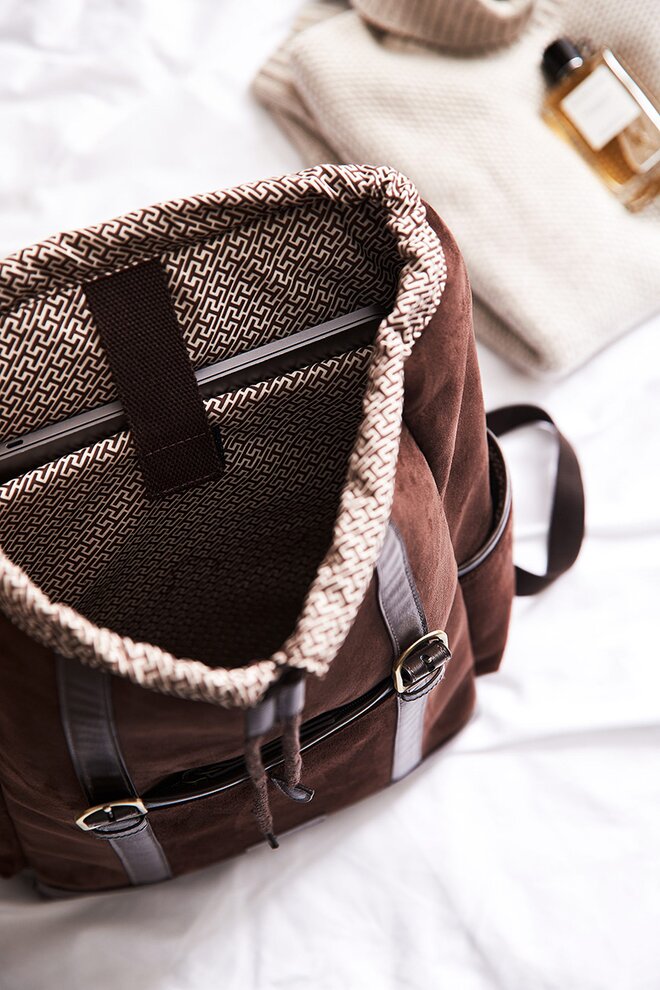 Elegant og romslig brun ryggsekk i vintage-stil med skinnstropper og mykt, teksturert fôr. Sekken ligger åpen på en hvit seng sammen med en kremfarget genser og en parfymeflaske, som gir en følelse av reise og luksus.